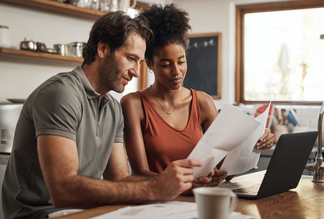 A couple at their kitchen counter reviewing paperwork together beside an open laptop.